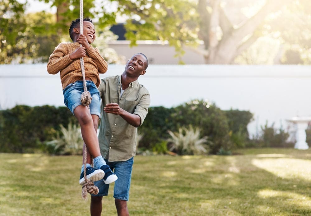 Father pushing son on a rope swing in a sunny backyard.