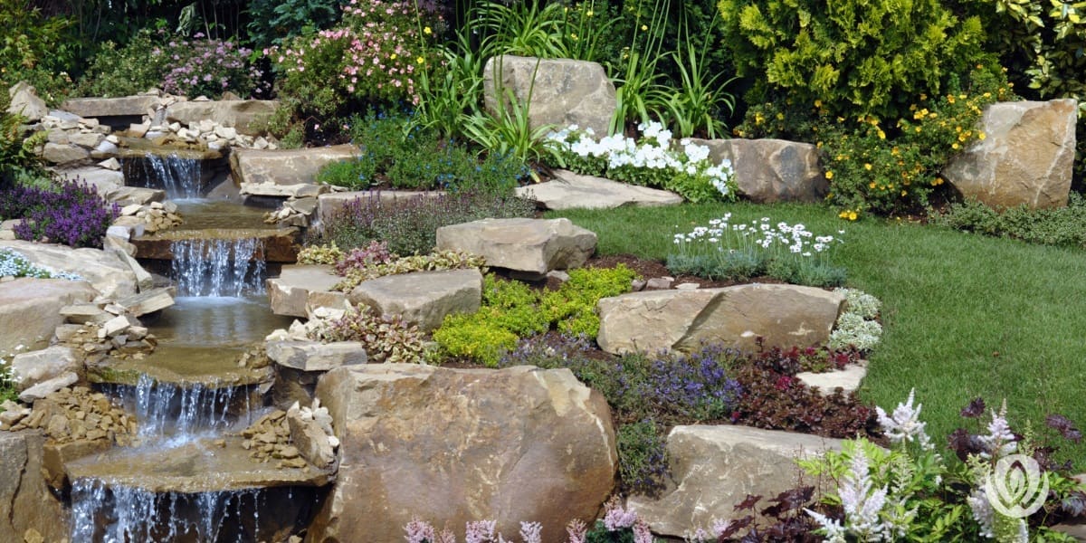 Serene backyard water feature with natural stone, blooming flowers, and surrounding greenery creating a relaxing outdoor oasis